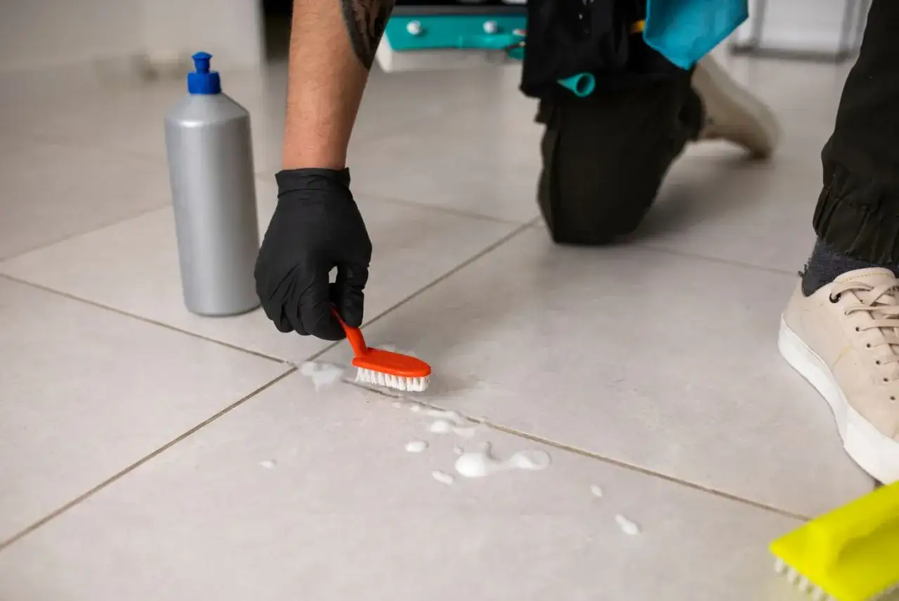 Main tenant utilise une brosse pour nettoyer carrelage extérieur antidérapant. Une bouteille de produit nettoyant est à côté.