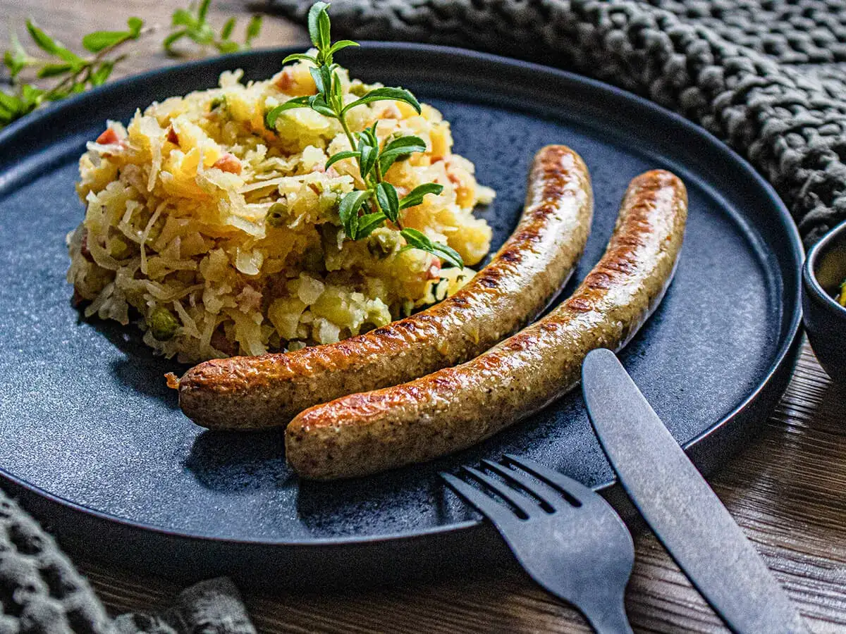 Zwei goldbraune Bratwürste im Airfryer, serviert mit deftigem Kartoffel-Krautsalat auf einem dunklen Teller.