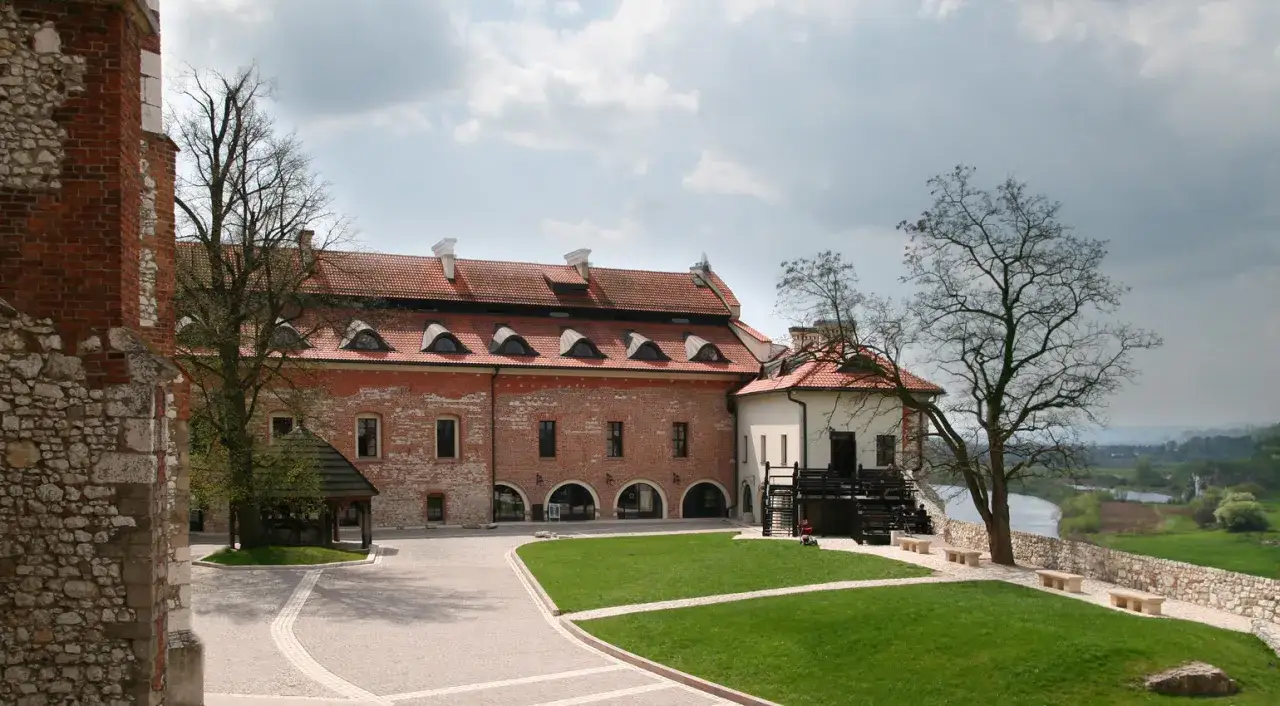 Kamienny dziedziniec z zielonym trawnikiem i budynkami przypominającymi klasztor dla świeckich. W oddali rzeka i pagórkowaty krajobraz.