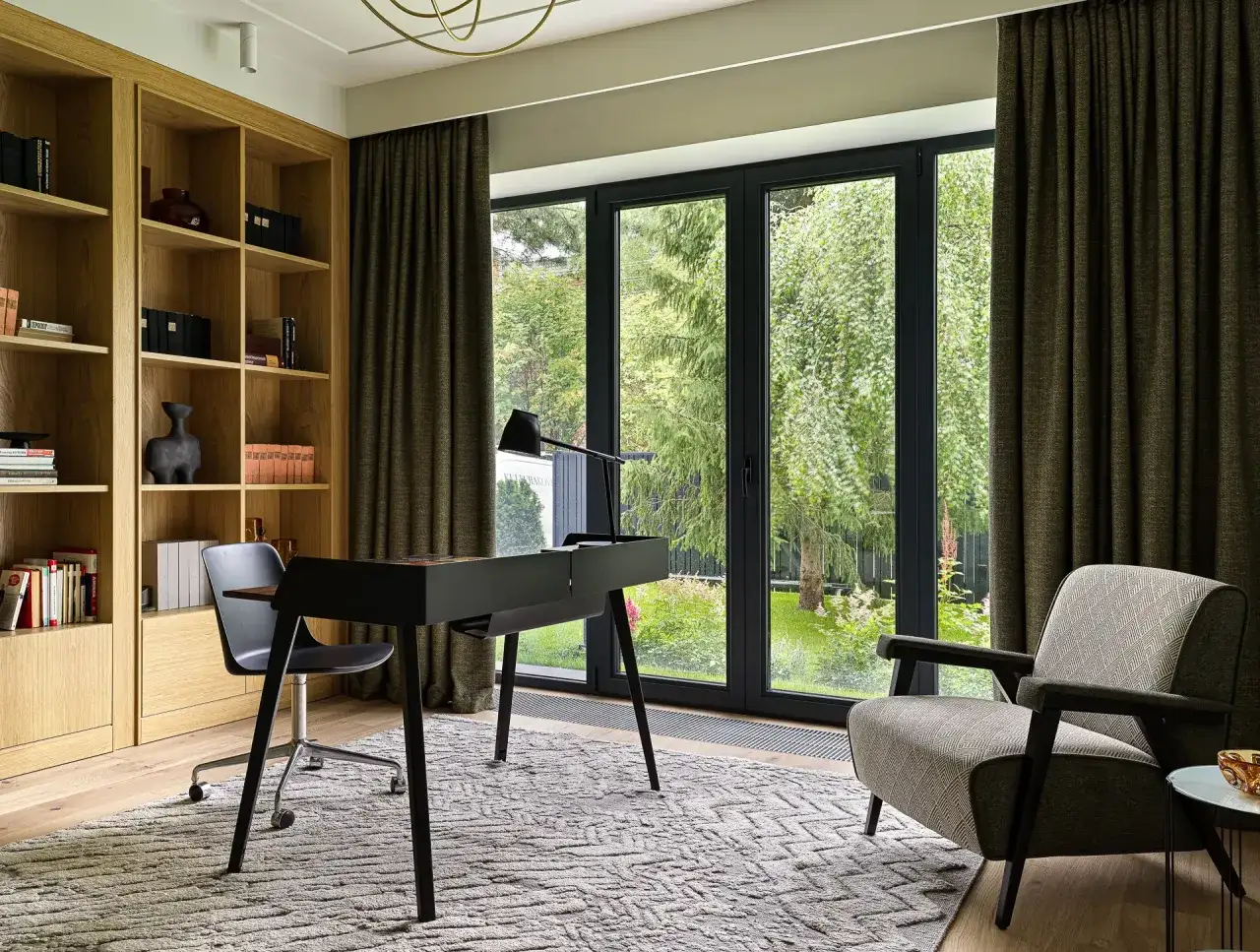 Acogedora habitaci&oacute;n despacho en casa con estanter&iacute;as de madera, escritorio negro, silla ergon&oacute;mica y sill&oacute;n. Grandes ventanales con vistas al jard&iacute;n.