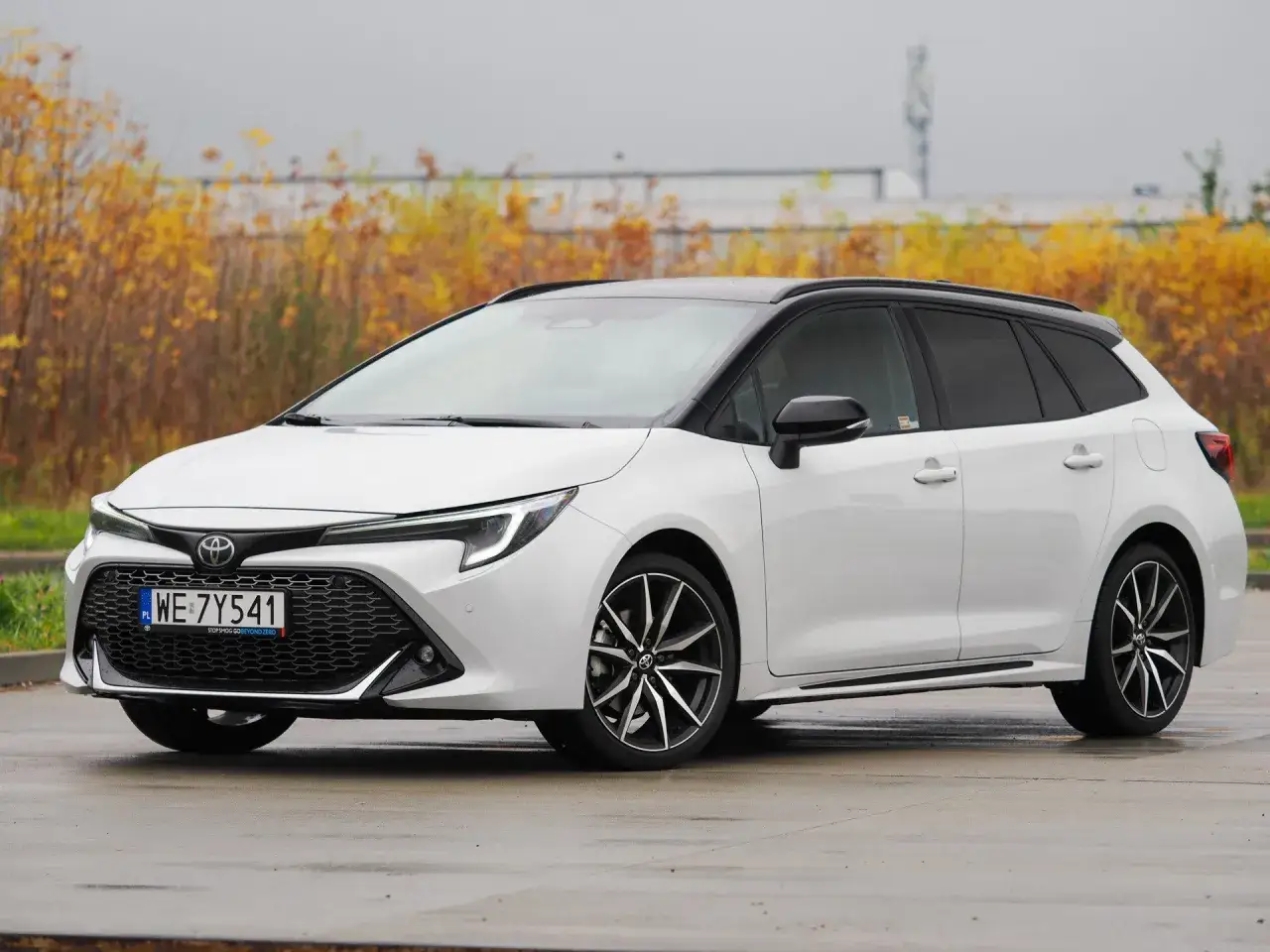Biała Toyota Corolla Hybrid w wersji kombi, z czarnym dachem, zaparkowana na mokrym asfalcie. W tle jesienne krzewy.