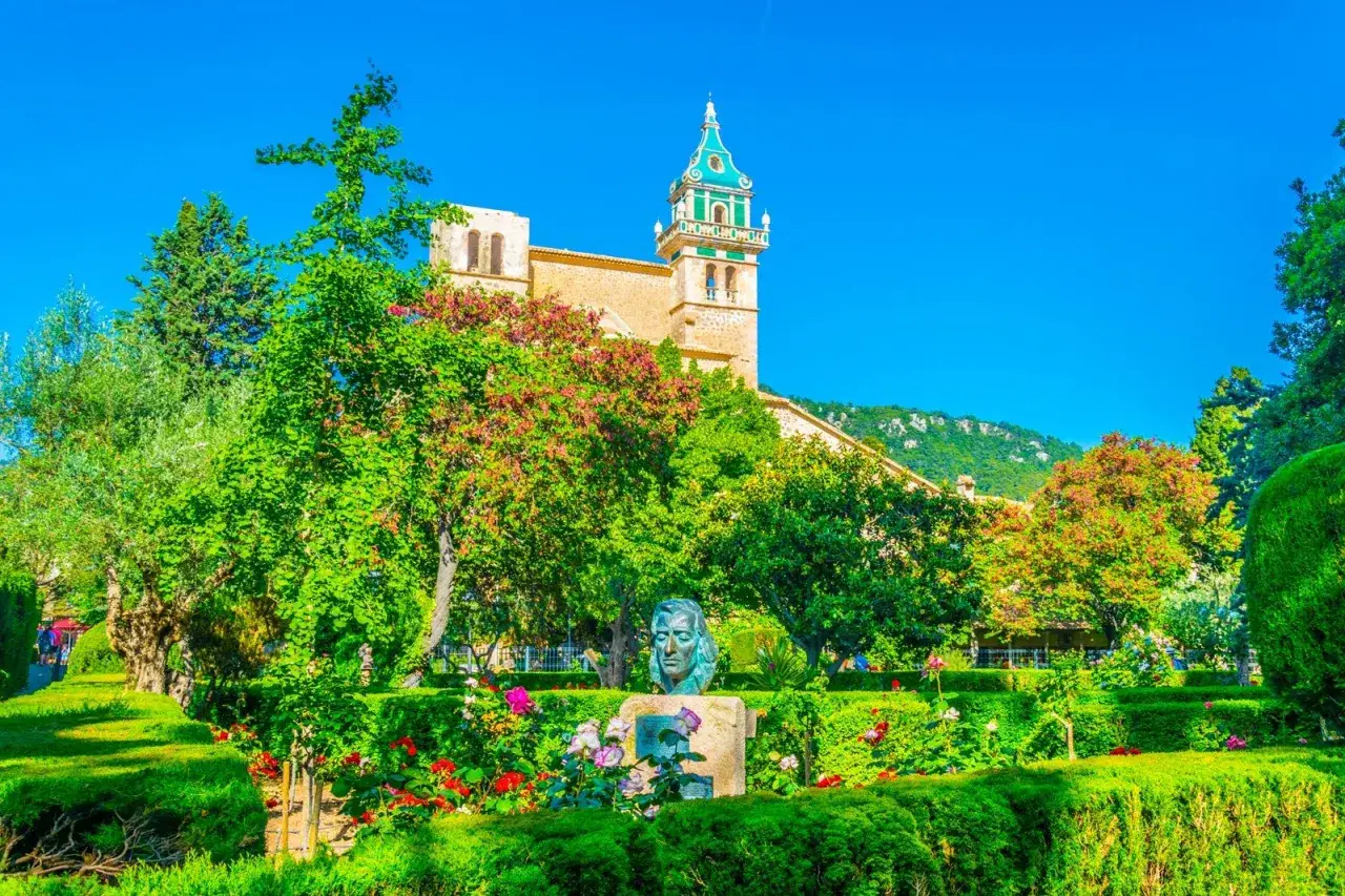 A lush garden with a bust leads to the historic Carthusian monastery, its bell tower reaching towards a clear blue sky.