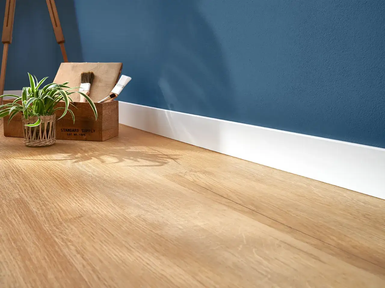 Un angolo di casa con un pratico **pavimento laminato** effetto legno, una pianta e una cassetta con pennelli.