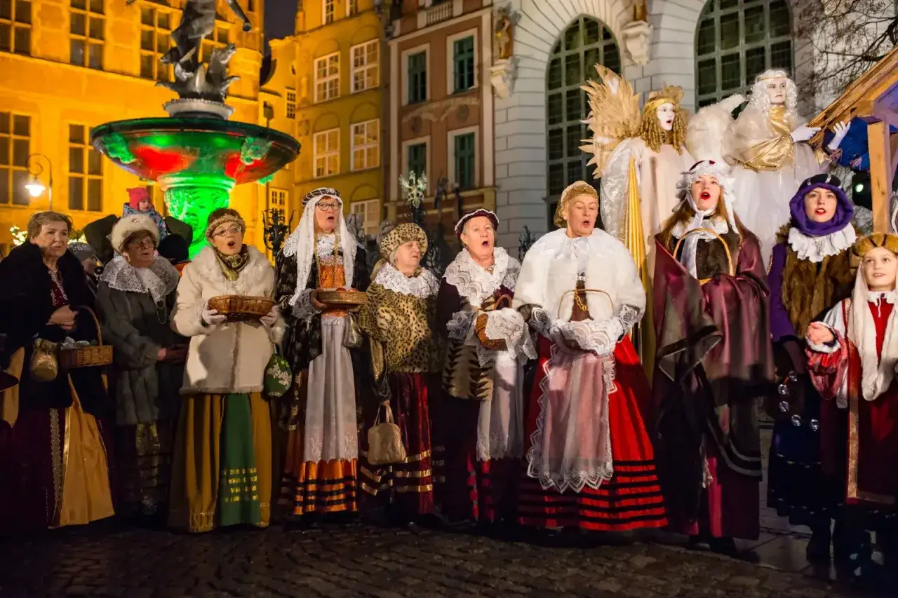 Grupa osób w strojach historycznych śpiewa, co oznacza słowo kolęda. W tle fontanna i kamienice.
