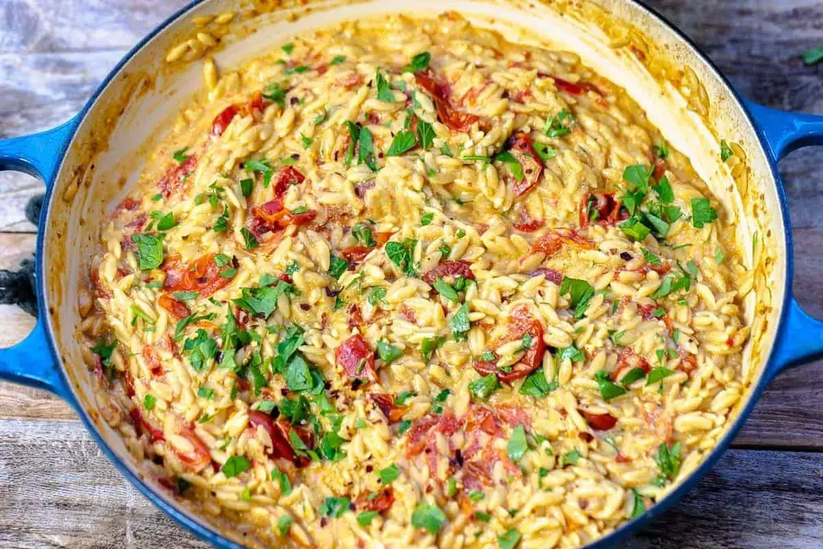 Cremiges Orzo-Rezept mit sonnengetrockneten Tomaten und frischer Petersilie, serviert in einem blauen Gusseisentopf.