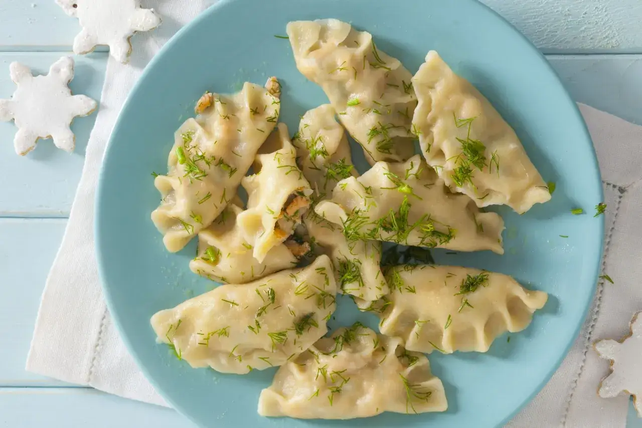 Pyszne pierogi z łososiem, posypane świeżym koperkiem, na jasnoniebieskim talerzu. Obok świąteczne ciasteczka w kształcie płatków śniegu.