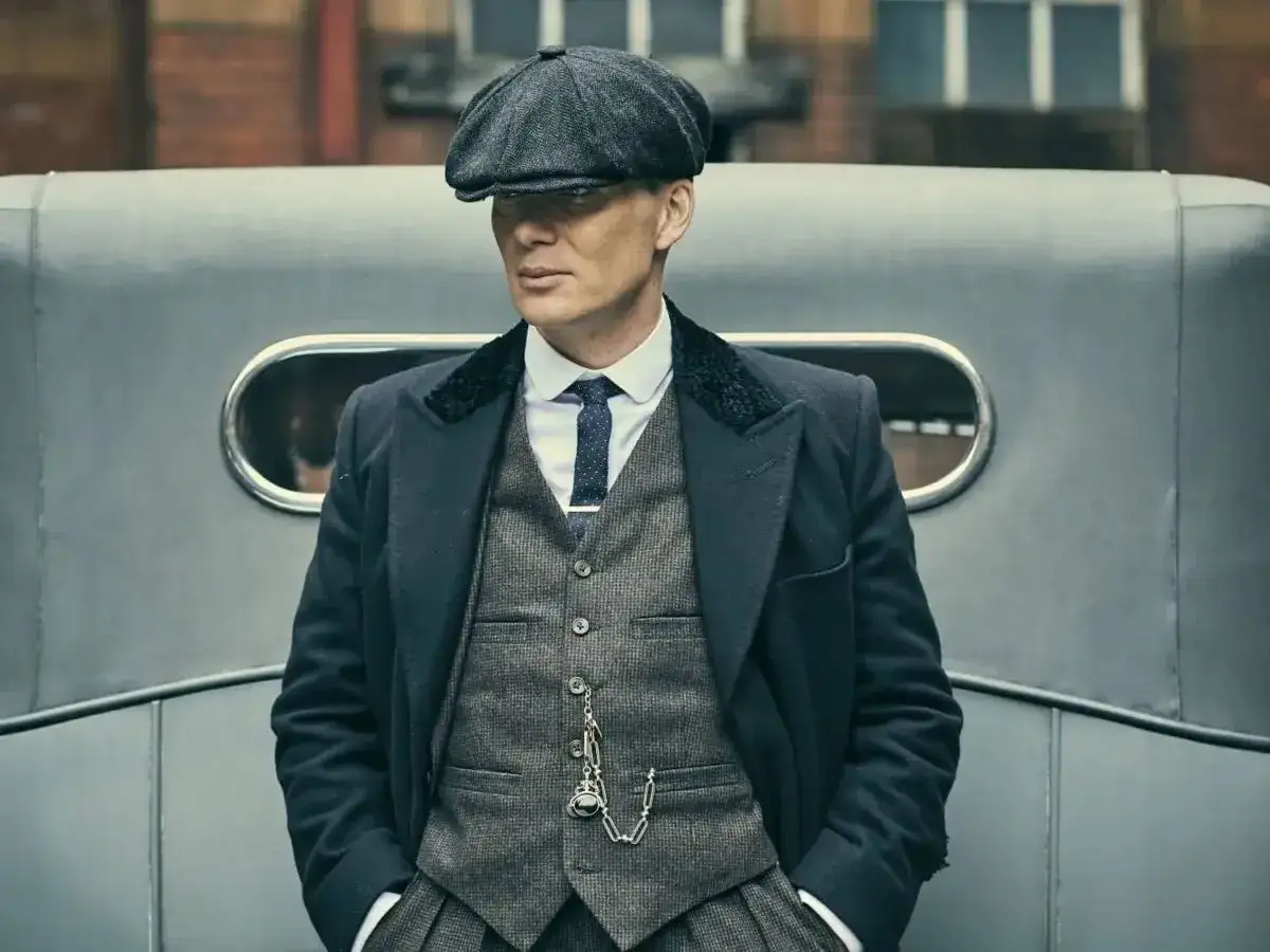 Tommy Shelby, con il suo iconico berretto e abito elegante, posa con aria pensierosa accanto a un'auto d'epoca.