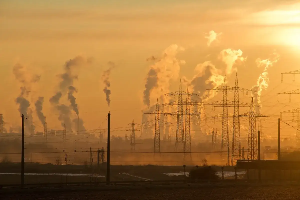Siluetas de chimeneas industriales emitiendo humo, creando una atmósfera de nox contaminante. Torres eléctricas se alzan contra el cielo anaranjado del atardecer.