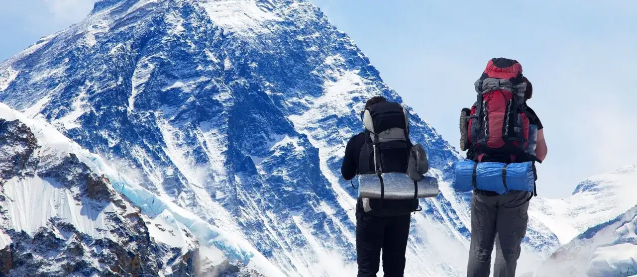 Dw&oacute;ch turyst&oacute;w z plecakami stoi przed majestatycznym, ośnieżonym szczytem. To właśnie tam leży Mount Everest, najwyższa g&oacute;ra świata.