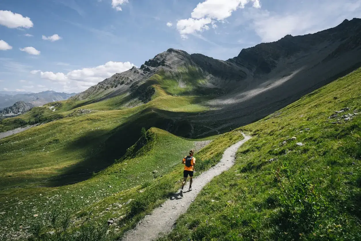 Biegacz na szlaku w Alpach, pokonujący trasę UTMB Mont Blanc. Zielone zbocza górskie i skaliste szczyty w tle.