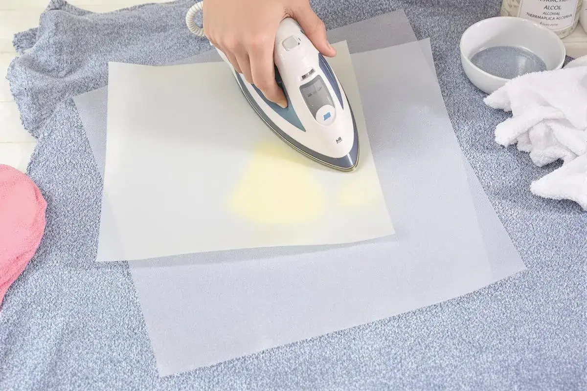 Utilisation d'un fer à repasser pour enlever colle thermocollant sur tissu, avec du papier sulfurisé pour protéger.