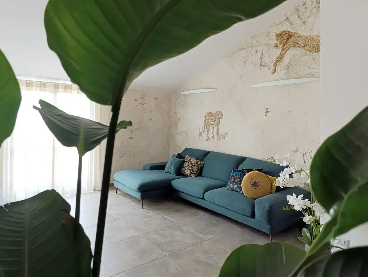 Divano turchese in un salotto con carta da parati a tema giungla e piante rigogliose, per un perfetto **stile jungle chic**.