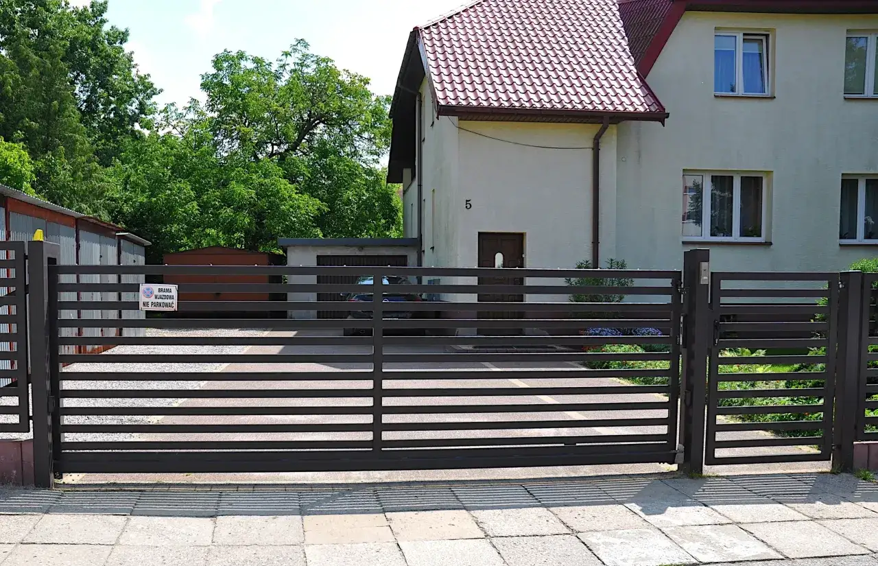 Nowoczesne ogrodzenie działki z bramą wjazdową. Na bramie widoczny znak zakazu parkowania, co może sugerować lokalne przepisy.