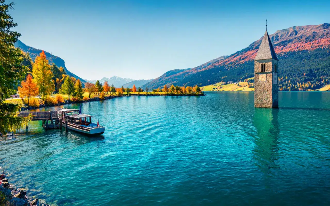Torre sommersa nel lago di Resia, un luogo magico che ispira tante curon recensioni. Autunno con colori vivaci e un battello attraccato.