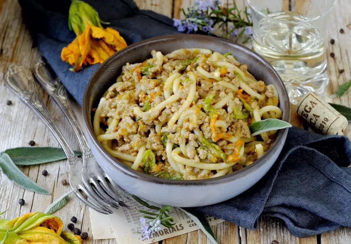 Pici al rag&ugrave; con fiori di zucca, un piatto rustico e saporito, servito in una ciotola su un tavolo di legno.