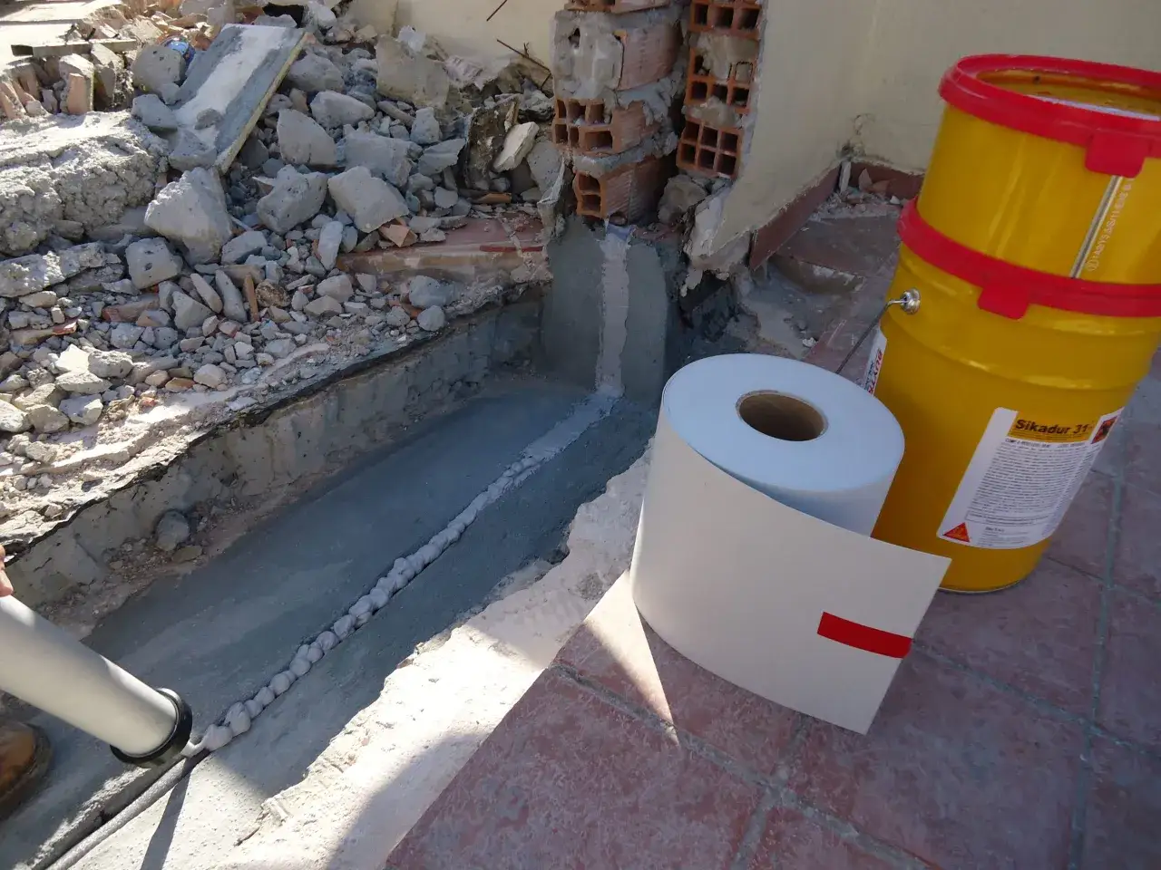 Aplicación de sellador para juntas de dilatación en obra. Se observa un rollo de material, un cubo amarillo de Sikadur 31 y una pistola aplicadora.