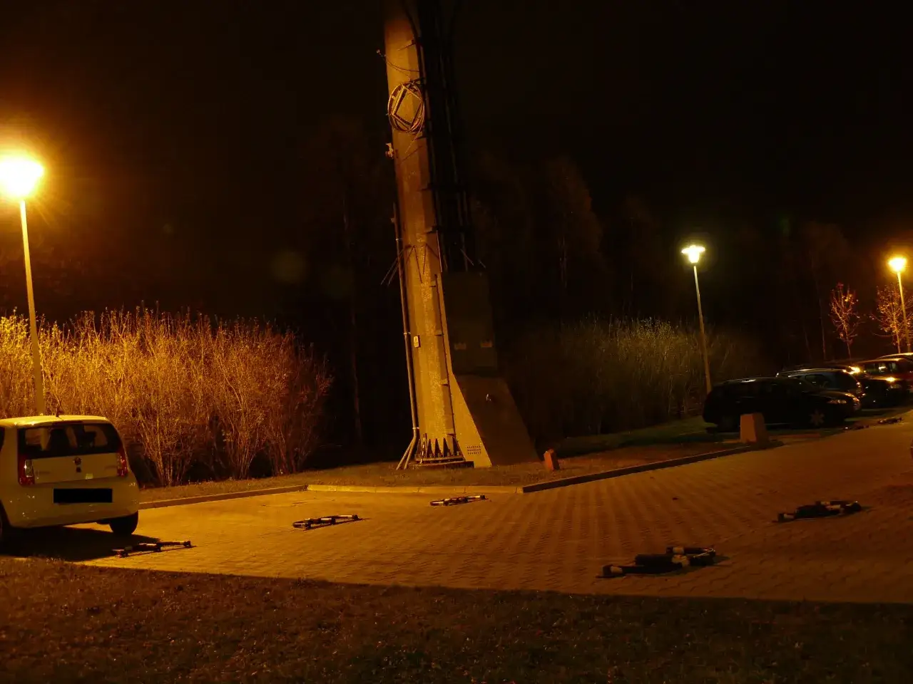 Nocny parking oświetlony lampą sodową, która nadaje żółtawy odcień. W tle widać drzewa i słup energetyczny.