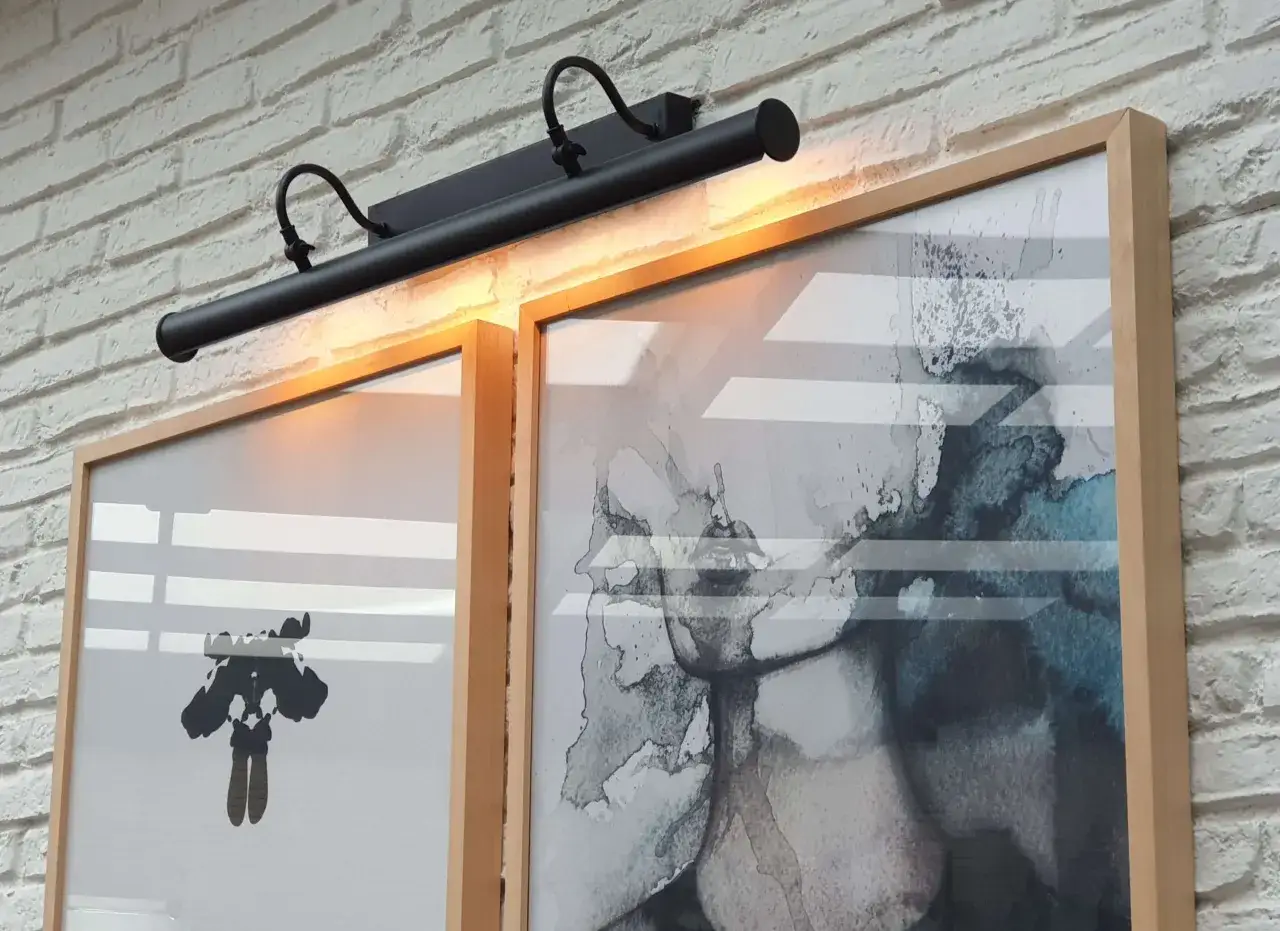 Aplique de pared negro ilumina dos cuadros modernos con marcos de madera. Uno tiene una figura abstracta en negro, el otro, acuarelas en tonos grises y azules.