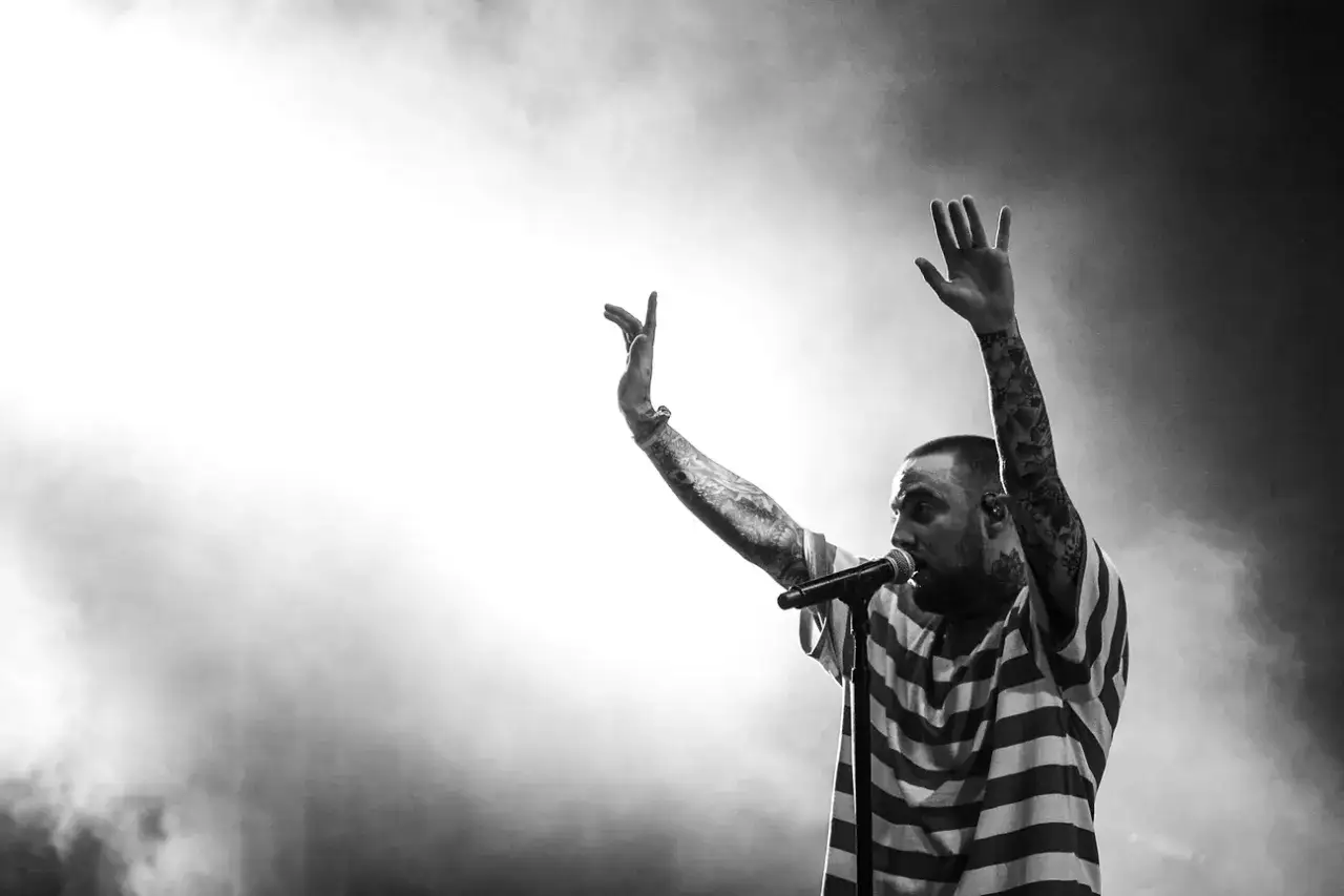 Mac Miller z podniesionymi rękami, śpiewający do mikrofonu. Jego śmierć wciąż porusza fan&oacute;w.