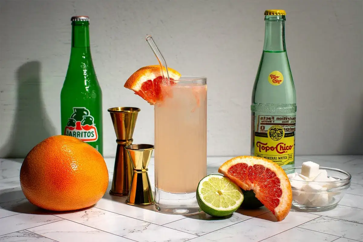 Paloma przepis: świeży grejpfrut, limonka, kostki cukru, butelka Jarritos i Topo Chico, złote miarki barmańskie i gotowy drink.