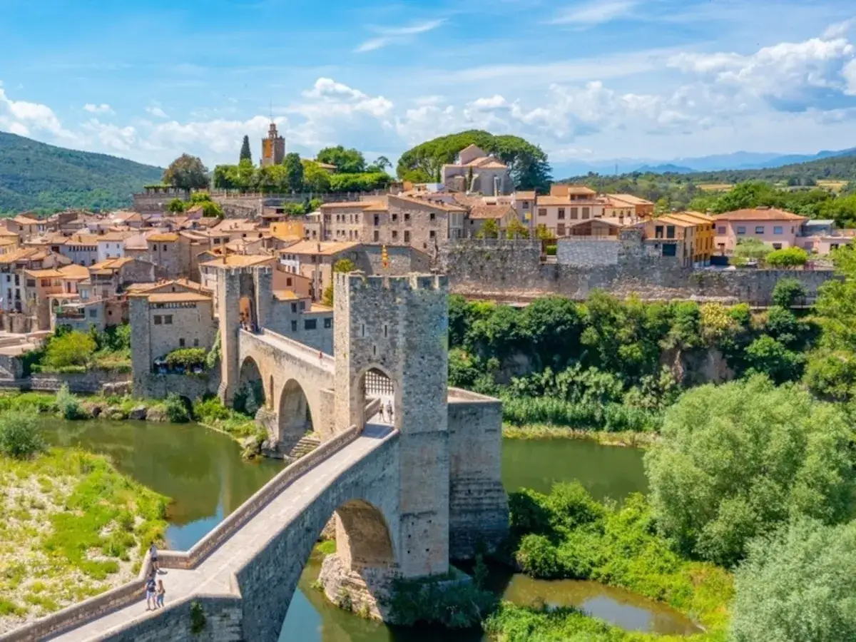 Puente medieval y casas de piedra en uno de los pueblos de Girona, con un río verde y vegetación exuberante.