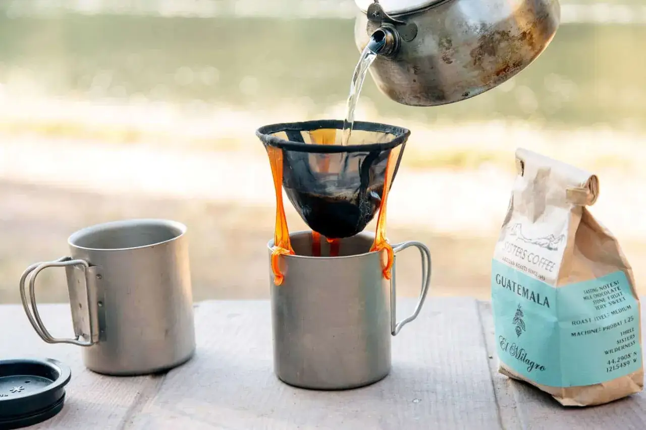 Hei&szlig;es Wasser flie&szlig;t in einen Filterhalter, um Kaffee zu kochen. Camping-Tassen und Kaffeepackung stehen bereit.