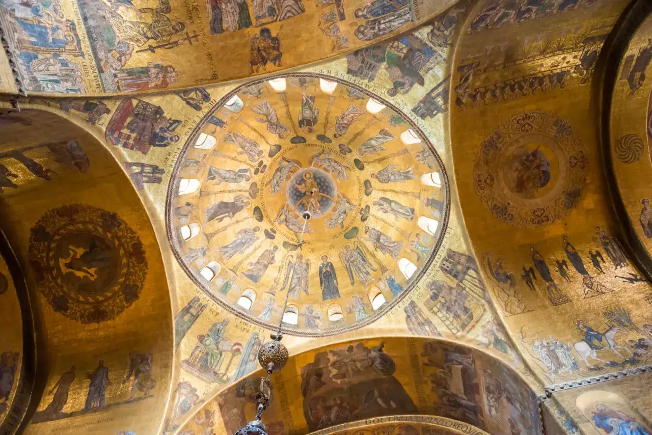 Mosaici dorati che ricoprono le volte di una chiesa, un esempio di **famous art in Venice**. Scene bibliche e figure sacre brillano sotto la luce.
