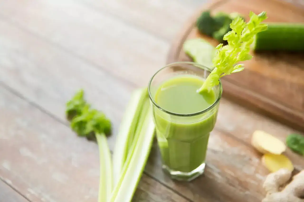 Grüner Selleriesaft im Glas, ideal für eine Sellerie Leberreinigung. Frische Zutaten wie Sellerie, Gurke und Ingwer liegen daneben.