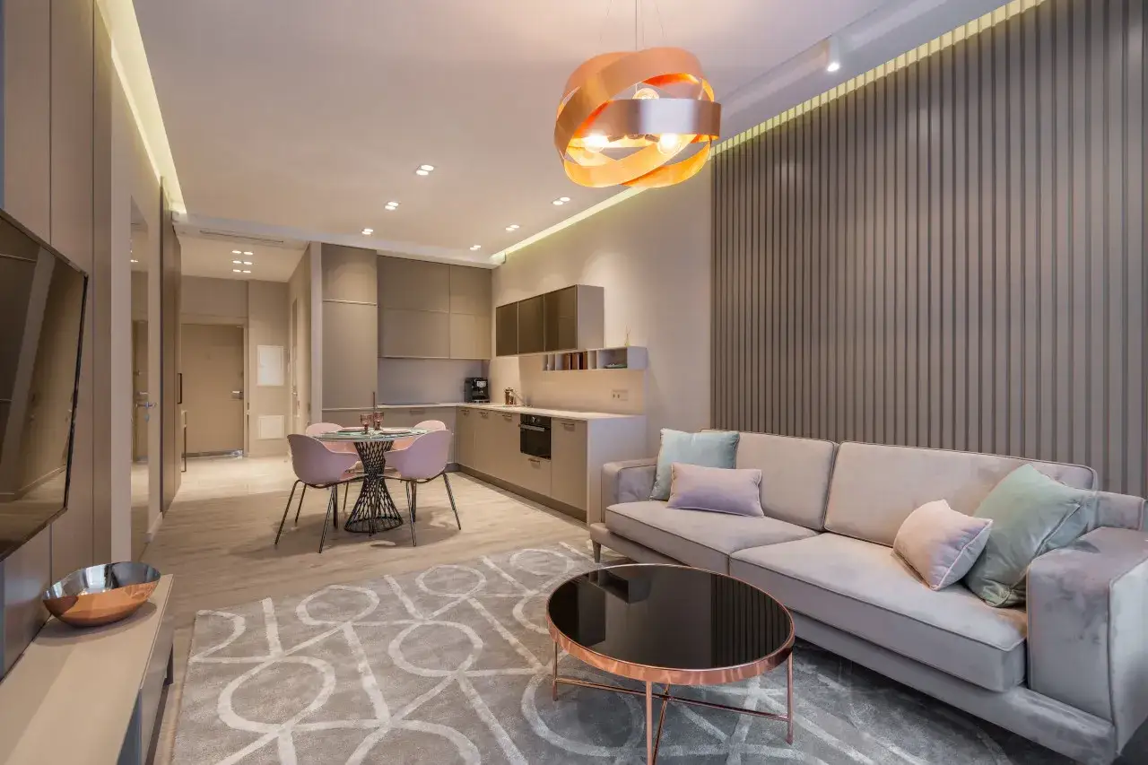 Nowoczesny salon 20m2 aranżacja: szara sofa, okrągły stolik, różowe krzesła przy stole, kuchnia w zabudowie.