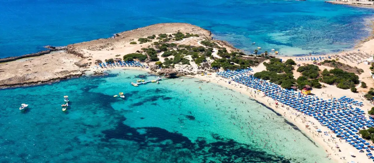 Słoneczny dzień na plaży w Ayia Napa. Tłumy ludzi odpoczywają pod niebieskimi parasolami, a w krystalicznie czystej wodzie pływają łodzie i kąpią się plażowicze.