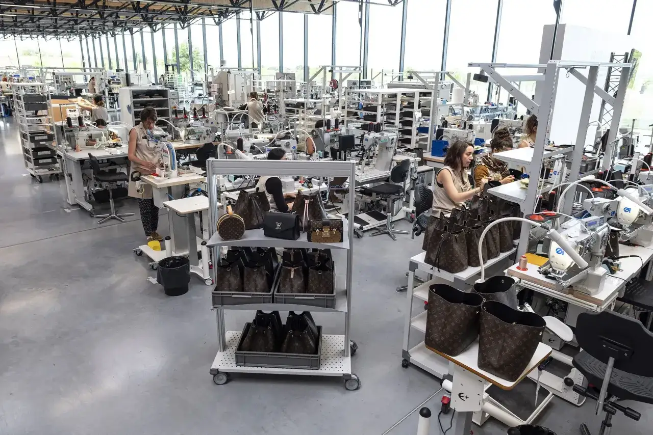 Operai al lavoro nella fabbrica Louis Vuitton, tra macchine da cucire e borse iconiche.