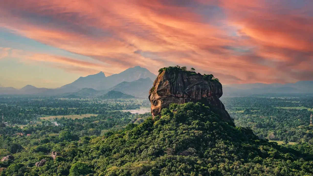 Sigiriya, Sri Lanka co warto zobaczyć: majestatyczna skała z ruinami pałacu na szczycie, otoczona bujną zielenią i malowniczymi górami pod zachodzącym słońcem.