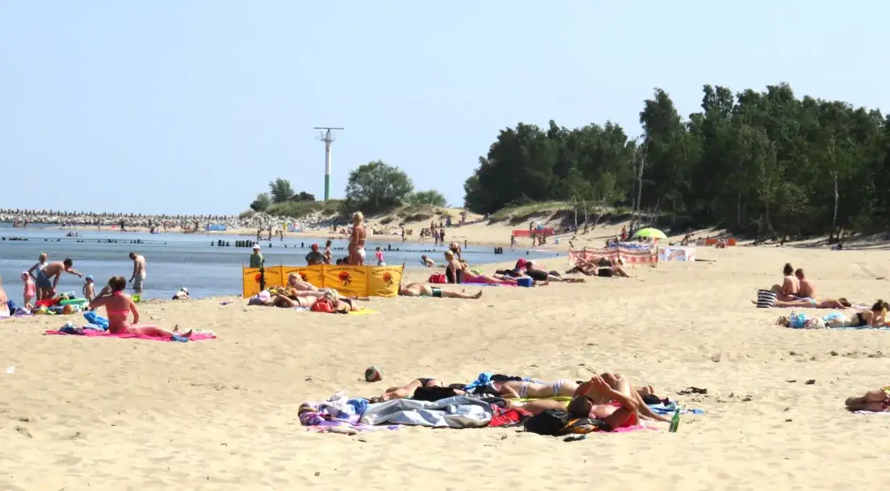 Słoneczny dzień na plaży w Górkach Zachodnich. Ludzie opalają się na piasku, niektórzy kąpią się w morzu. W tle widać fragment mariny.