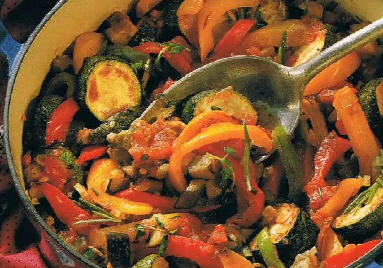 Kolorowy ratatuj przepis w garnku. Widoczne cukinia, papryka, pomidory i zioła. Łyżka zagłębia się w aromatyczne warzywa.