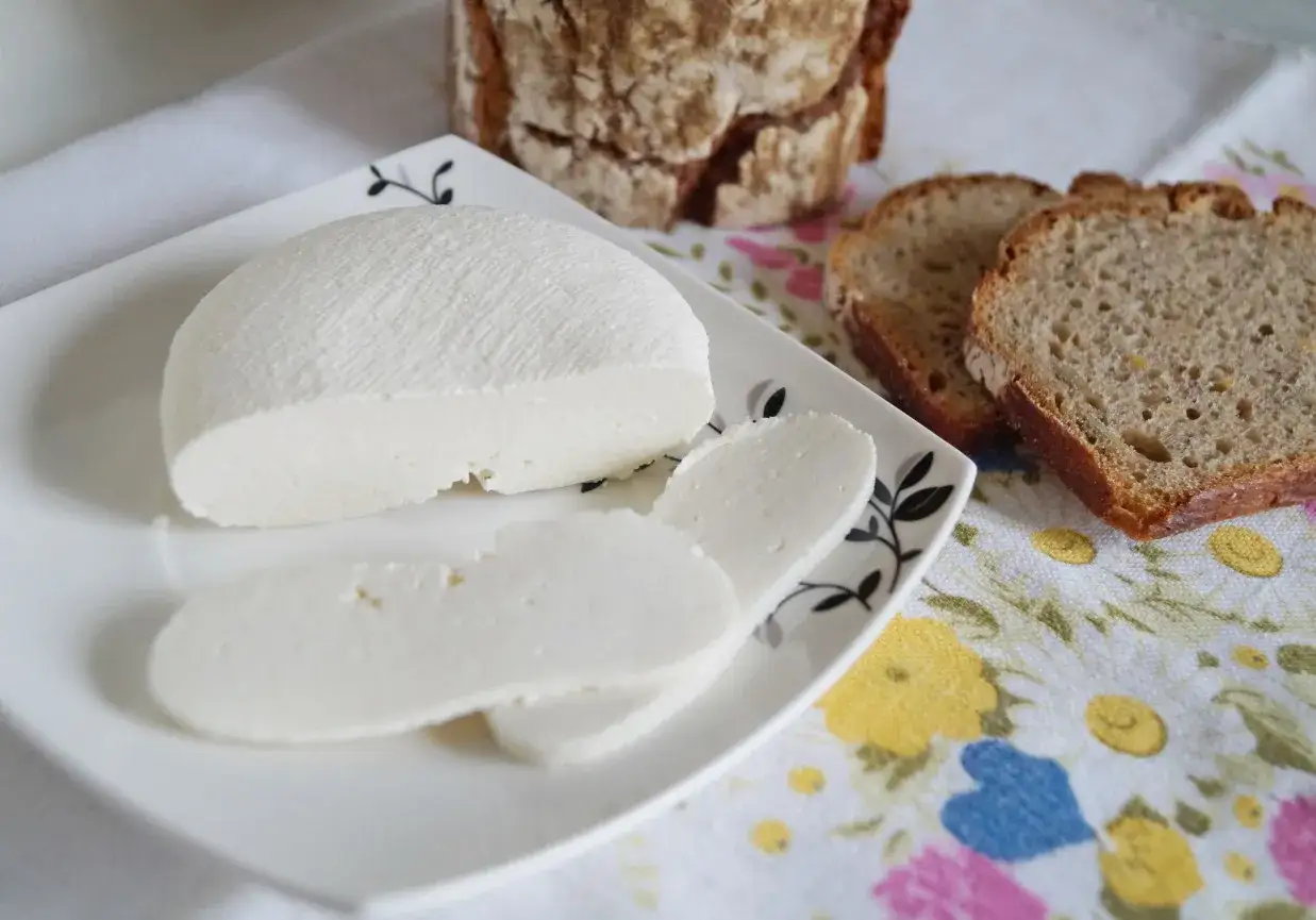 Domowy twaróg z mleka krowiego, pokrojony w plastry, obok kromek chleba. Idealny na śniadanie.