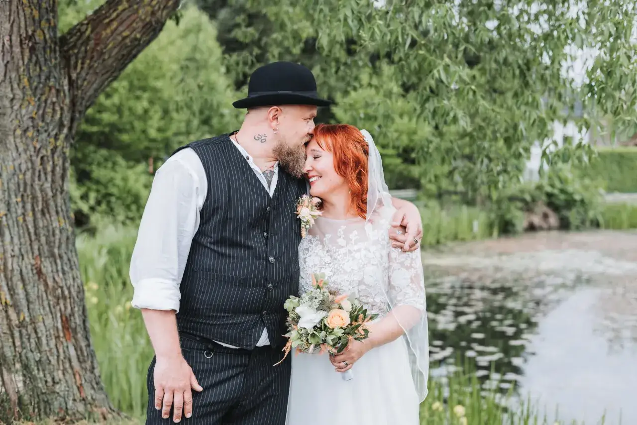 Ein Brautpaar umarmt sich liebevoll vor einem Teich. Der Bräutigam trägt einen Hut und eine Weste, die Braut ein Spitzenkleid. Ein schöner Spruch für die Ehe.