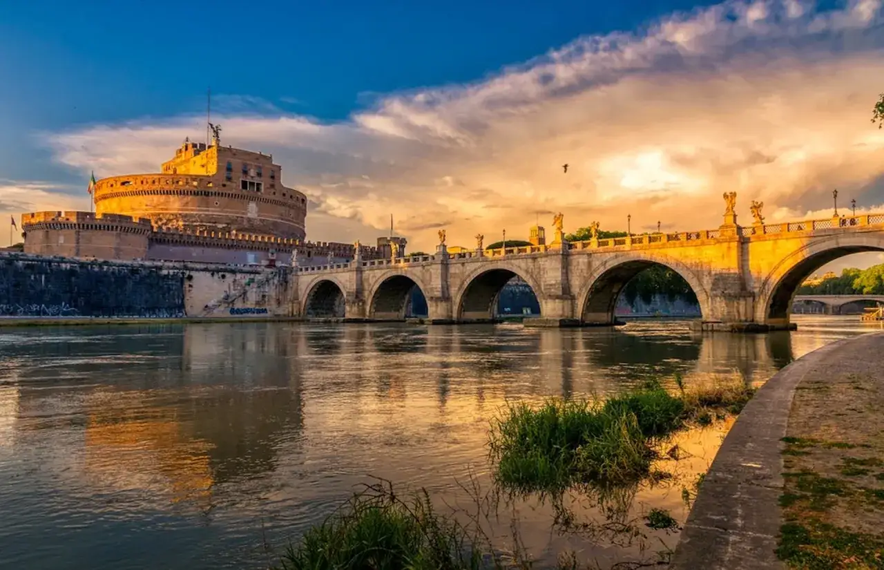 Le Castel Sant'Angelo se reflète dans le Tibre au coucher du soleil, sous un ciel aux teintes orangées.