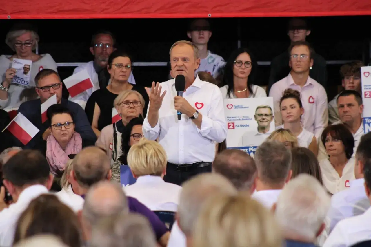 Tusk przemawia do tłumu, który trzyma polskie flagi. W tle widać plakaty wyborcze.