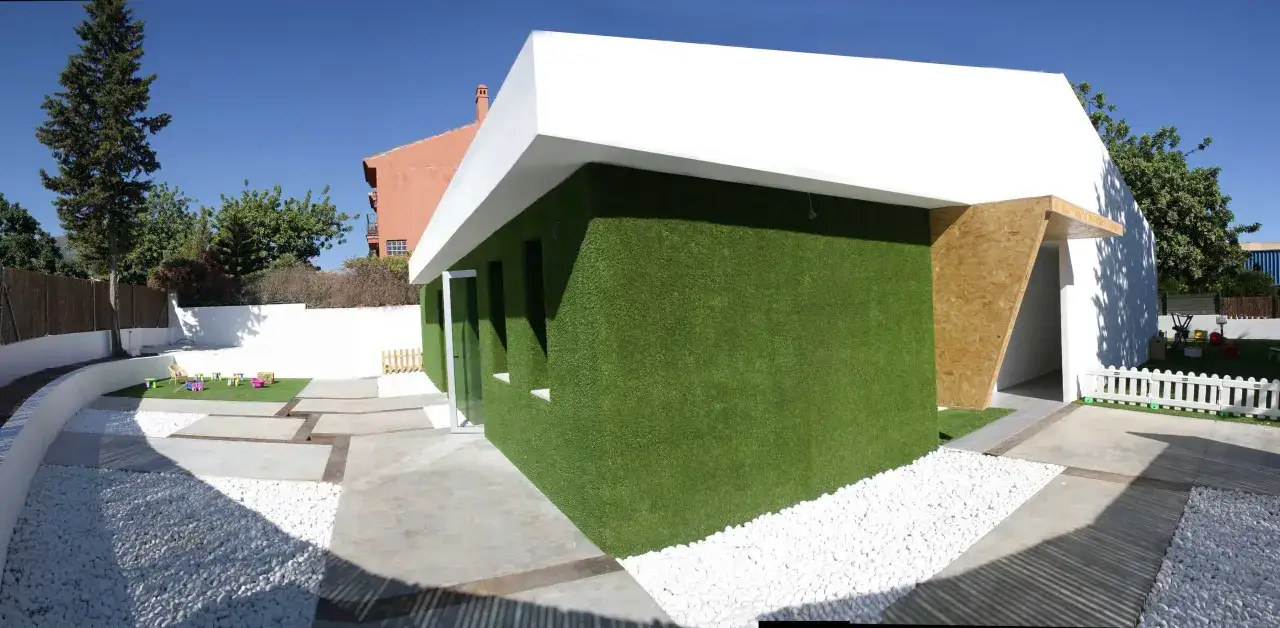 Edificio moderno con paredes cubiertas de distintos tipos de césped artificial. Patio con grava blanca y zona de juegos.