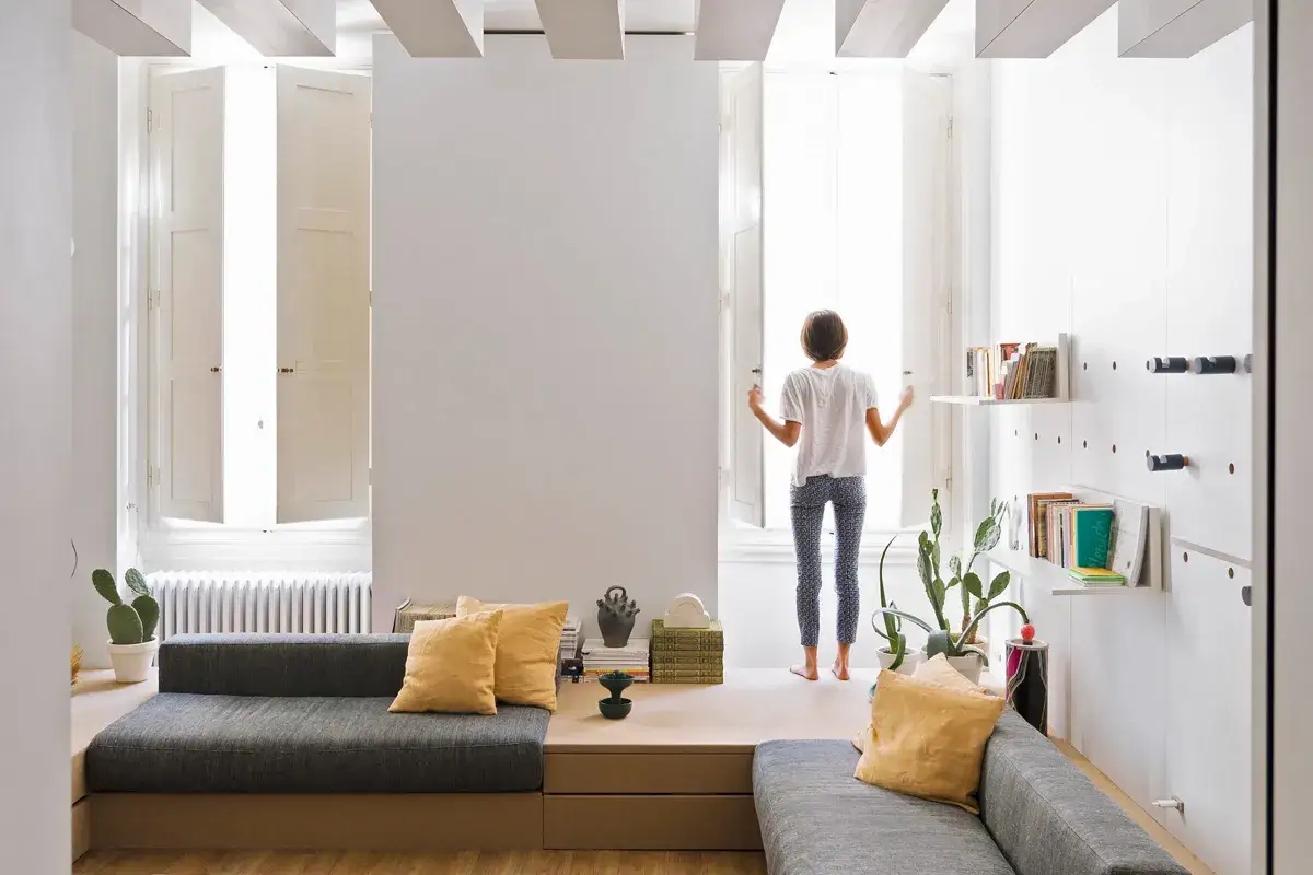 Arredare casa con poco: un salotto minimalista con divano angolare, librerie a muro e una persona che apre le finestre.