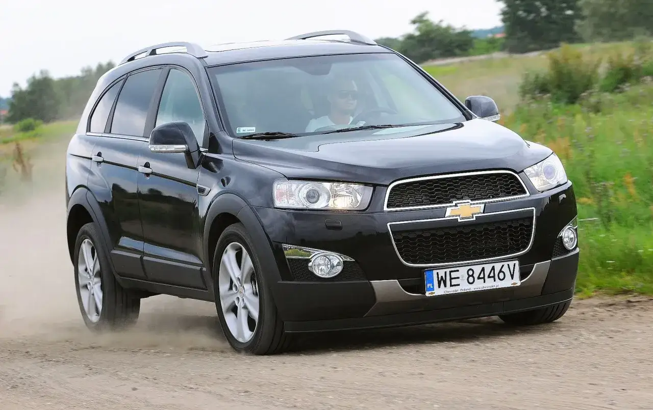 Czarny Chevrolet Captiva jedzie po piaszczystej drodze, wzbijając tumany kurzu. Kierowca w okularach przeciwsłonecznych prowadzi auto przez zielone pola.