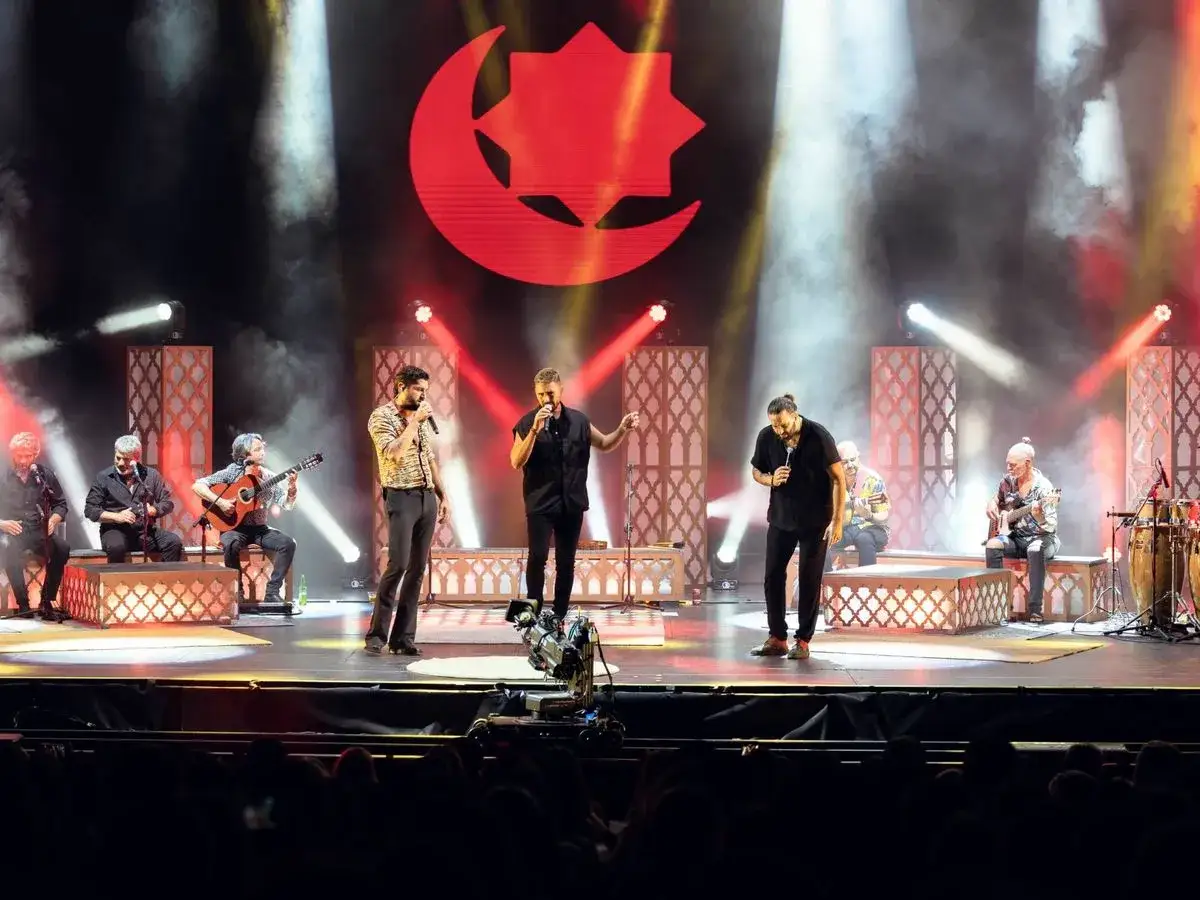 Artistas en un escenario con un gran logo rojo de luna y estrella. Un grupo interpreta m&uacute;sica con guitarras y percusi&oacute;n, mientras tres cantantes lideran la actuaci&oacute;n. El **fondo flamenco baile** crea una atm&oacute;sfera vibrante.