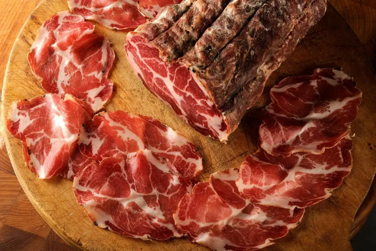 Fette di coppa su tagliere di legno, con un pezzo intero di coppa stagionata. Un assaggio di cos'è la coppa, un salume pregiato.