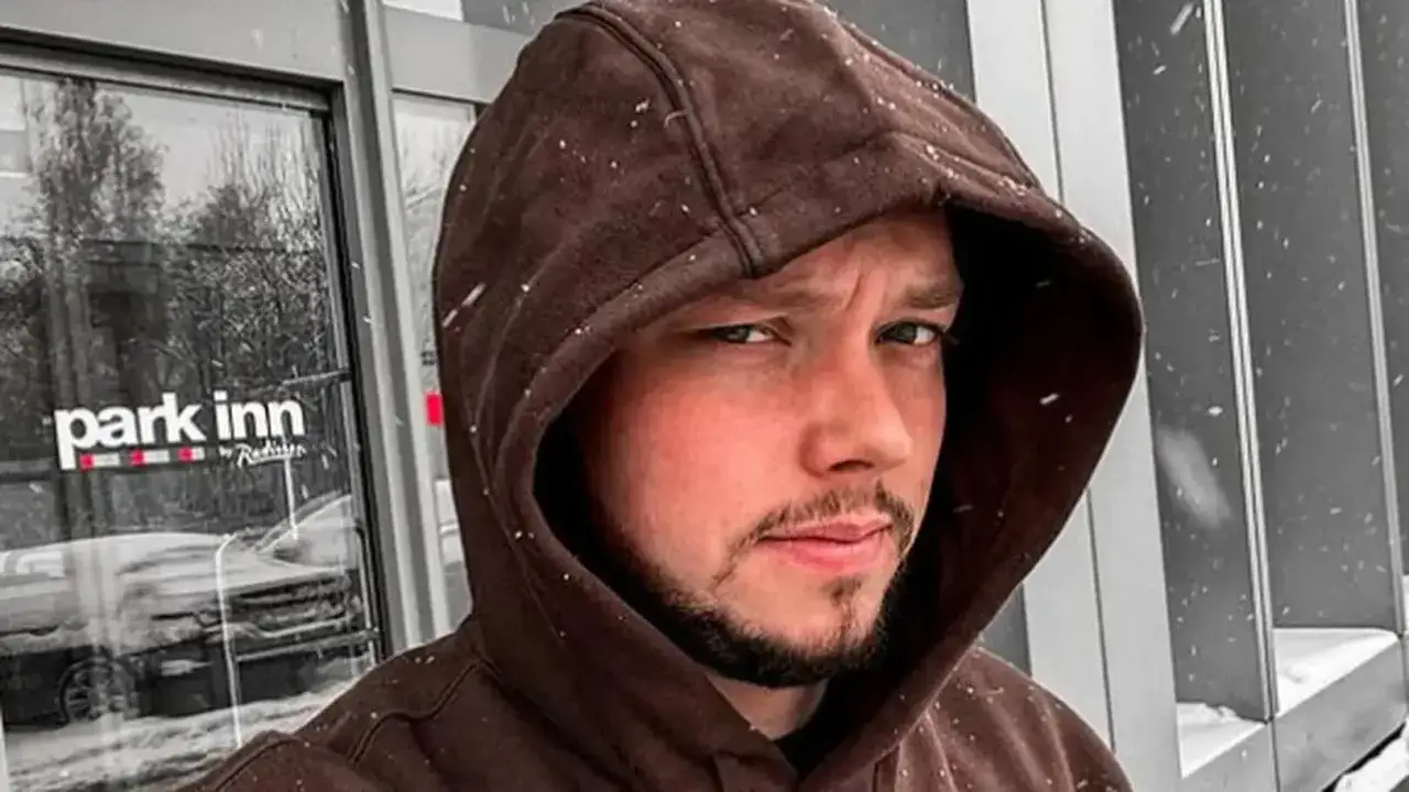 Młody mężczyzna w brązowej bluzie z kapturem, na tle drzwi hotelu Park Inn. Wokół pada śnieg, jakby to był księżulo.