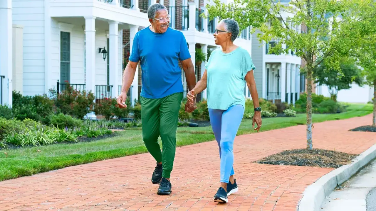 Pareja mayor pasea felizmente de la mano, disfrutando de un agradable **andar después de comer** por un sendero de ladrillos frente a casas blancas.