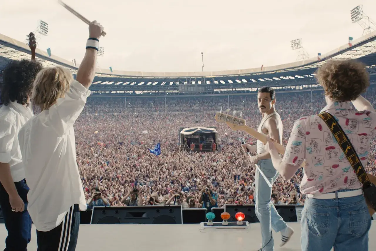 Zespół Queen na scenie stadionu, tłum szaleje, Freddie Mercury śpiewa. We will rock you!
