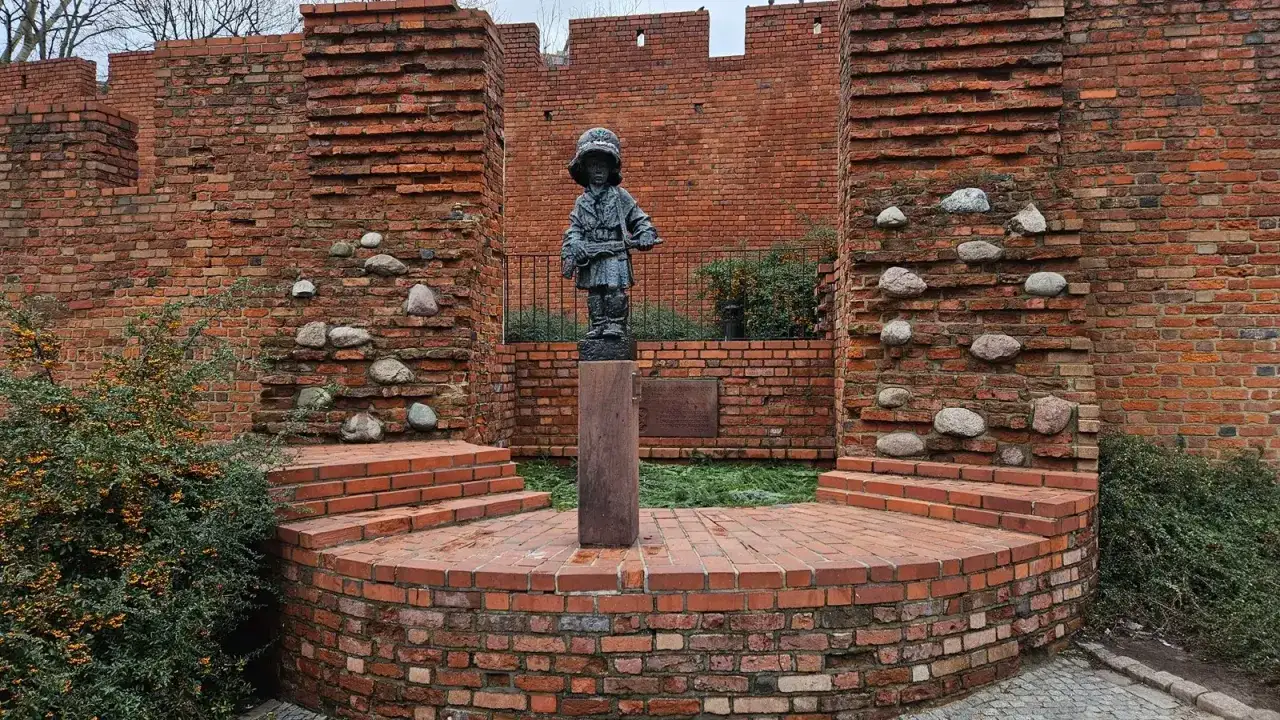 Pomnik Małego Powstańca, symbol wiersza o dzieciach walczących w powstaniu warszawskim, stoi na tle ceglanej fortecy.