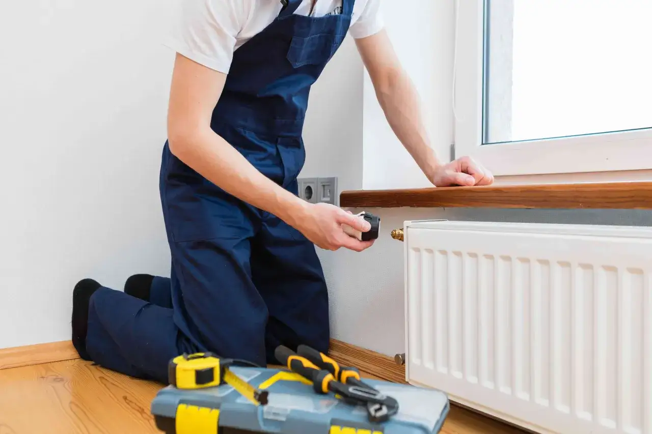 Instalator w granatowym kombinezonie pokazuje, jak zamontować grzejnik, trzymając termostat. Obok leży skrzynka z narzędziami.