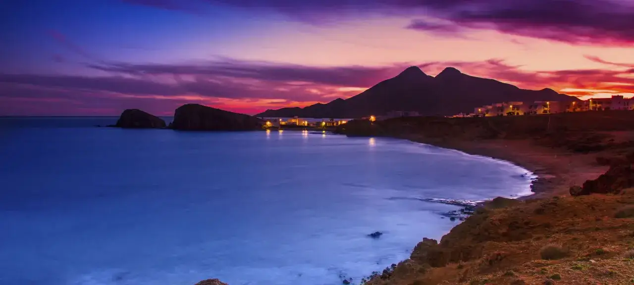 Atardecer sobre las playas vírgenes de Almería. El cielo se tiñe de morado y naranja, el mar azul acaricia la costa rocosa y las luces de un pequeño pueblo brillan al fondo.