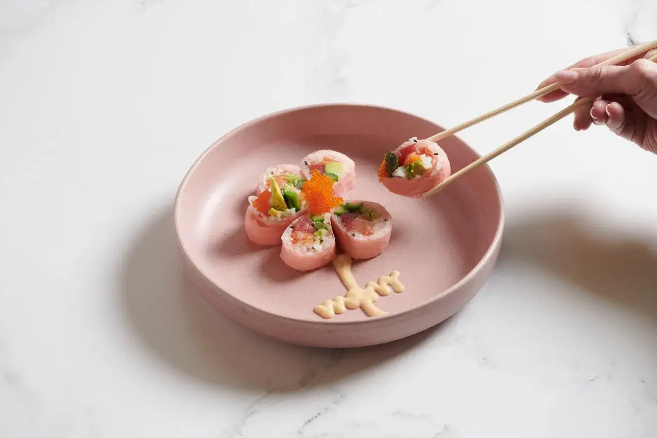 Różowe rolki sushi z łososiem i awokado, podane na talerzu. Pałeczki podnoszą jedną rolkę, pokazując, jak jeść sushi.
